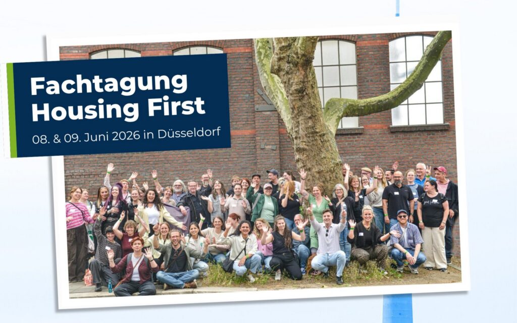 Eine große Gruppe von Menschen posiert und winkt vor einem Gebäude mit einem großen Baum, neben einem Schild für die Fachtagung „Housing First“ in Düsseldorf vom 8. bis 9. Juni 2026.