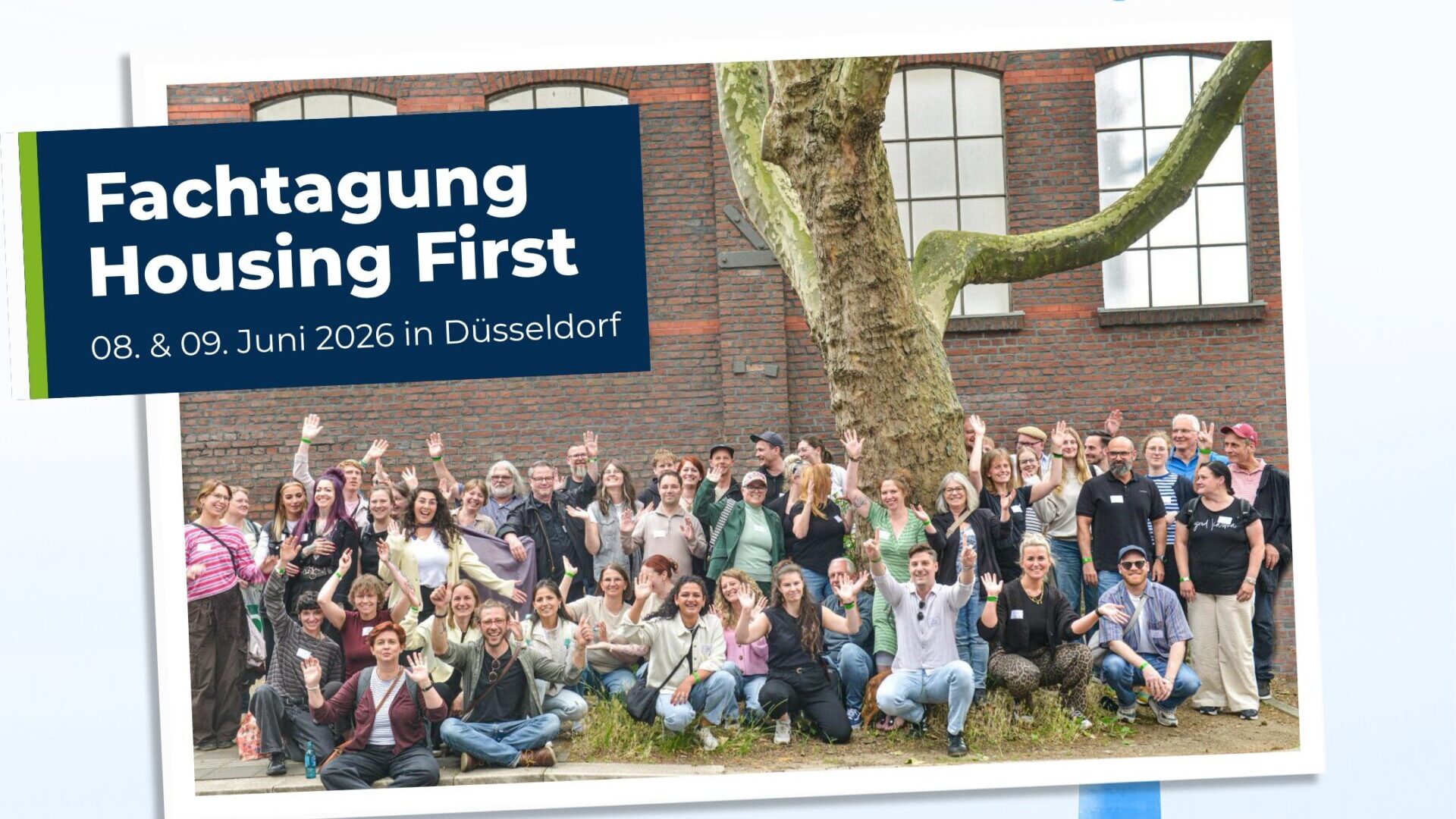 Eine große Gruppe von Menschen posiert und winkt vor einem Gebäude mit einem großen Baum, neben einem Schild für die Fachtagung „Housing First“ in Düsseldorf vom 8. bis 9. Juni 2026.