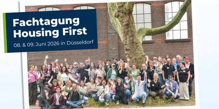 Eine große Gruppe von Menschen posiert und winkt vor einem Gebäude mit einem großen Baum, neben einem Schild für die Fachtagung „Housing First“ in Düsseldorf vom 8. bis 9. Juni 2026.