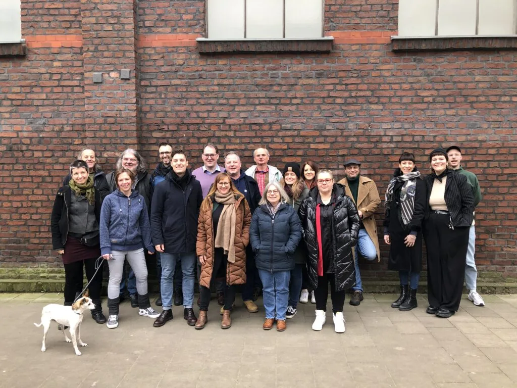 Eine Gruppe von 18 Erwachsenen und ein Hund stehen draußen vor einer roten Backsteinmauer. Die meisten tragen Winterjacken und Schals. Die Gruppe steht eng beieinander, und eine Person links hält die Leine des Hundes.