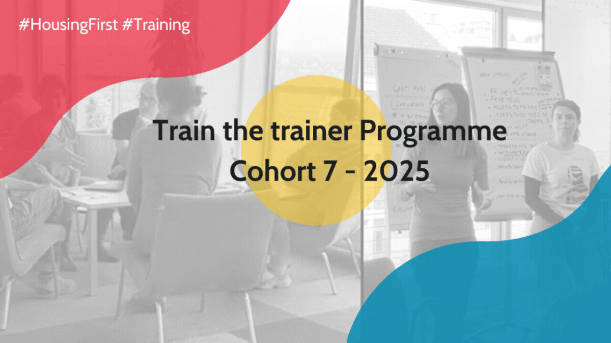 Eine Gruppe von Personen sitzt in einem Besprechungsraum, während eine Frau neben einem Flipchart steht und spricht. Textüberlagerung liest: "Train the trainer Programme Cohort 7 - 2025" mit #HousingFirst #Training.