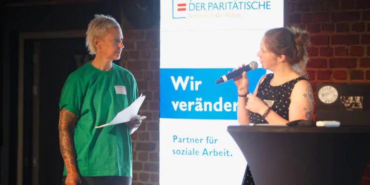Zwei Personen stehen mit Mikrofonen und Zetteln auf der Bühne vor einem Schild mit der Aufschrift „Wir verändern“ und „Partner für soziale Arbeit“.