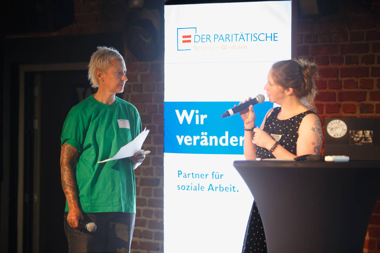 Zwei Personen stehen mit Mikrofonen und Zetteln auf der Bühne vor einem Schild mit der Aufschrift „Wir verändern“ und „Partner für soziale Arbeit“.