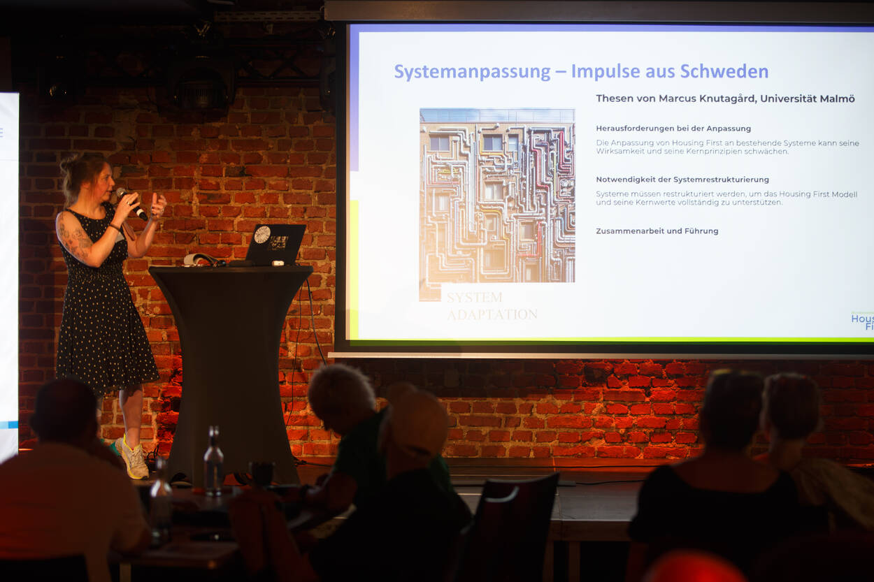 Eine Frau hält vor einem Publikum einen Vortrag über Systemanpassung; eine Folie mit Text und einer Grafik wird auf eine Backsteinmauer projiziert.