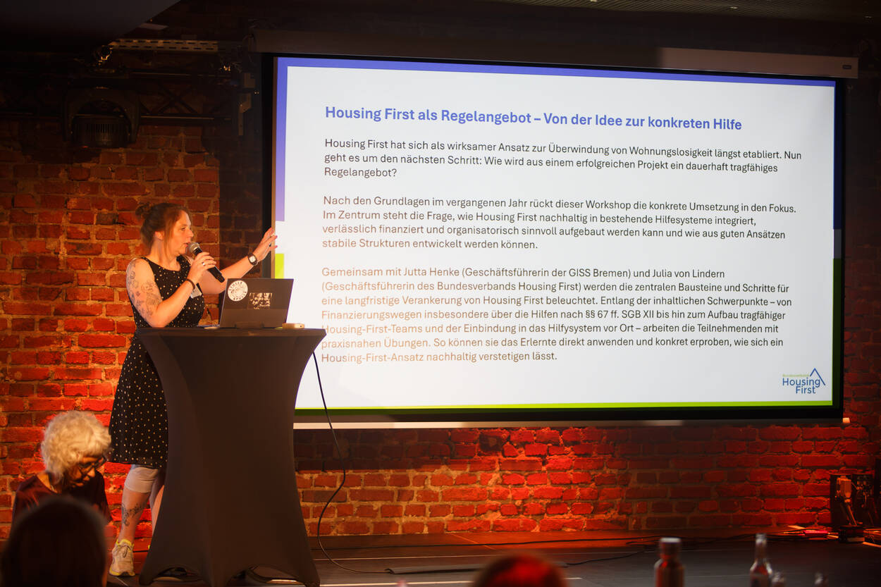 Eine Frau spricht an einem Rednerpult vor einer Backsteinmauer und präsentiert dem Publikum eine Folie über die „Housing First“-Initiative.